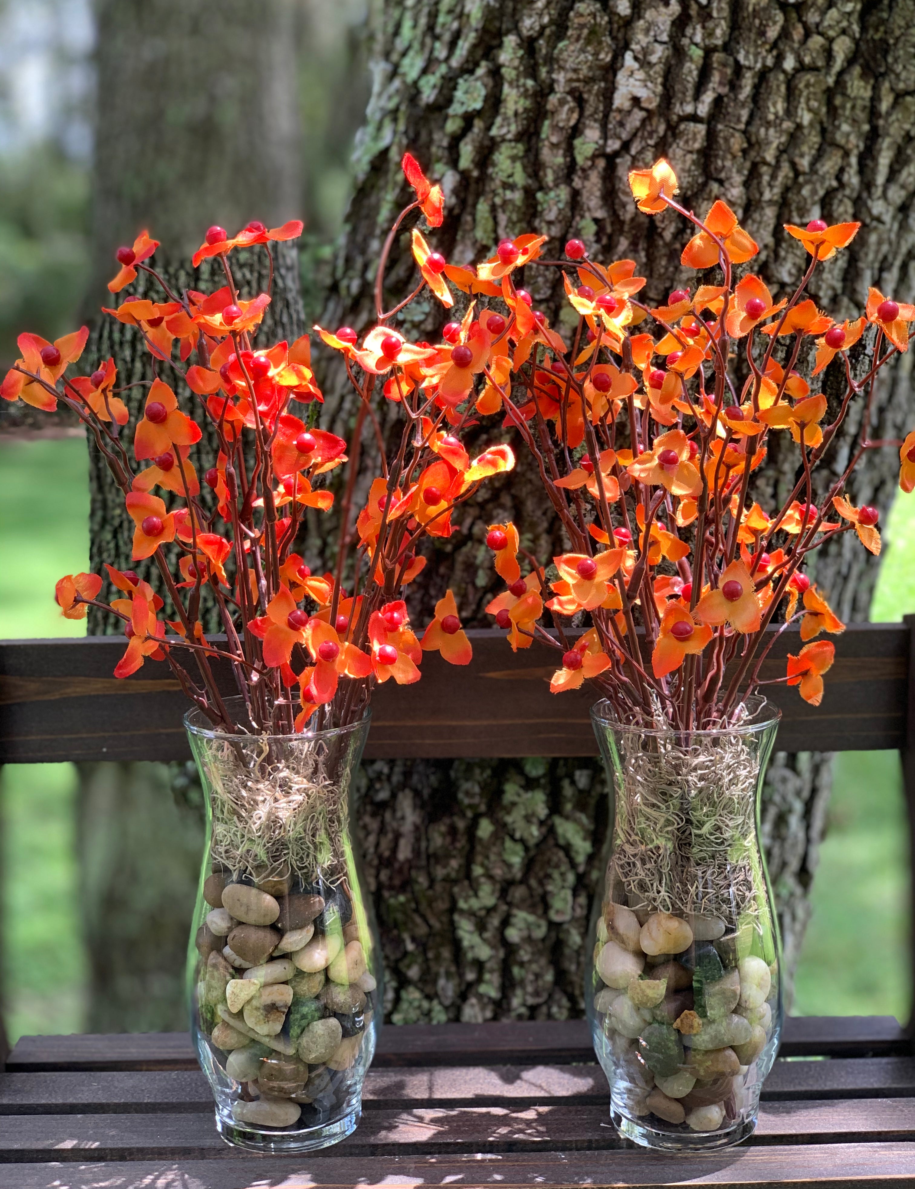 Bittersweet Floral Glass Vase shows an image of two vases together.  Each vase has tricolored earth tone rocks and tan moss with orange bittersweet artificial floral decor. Each sold separately.  