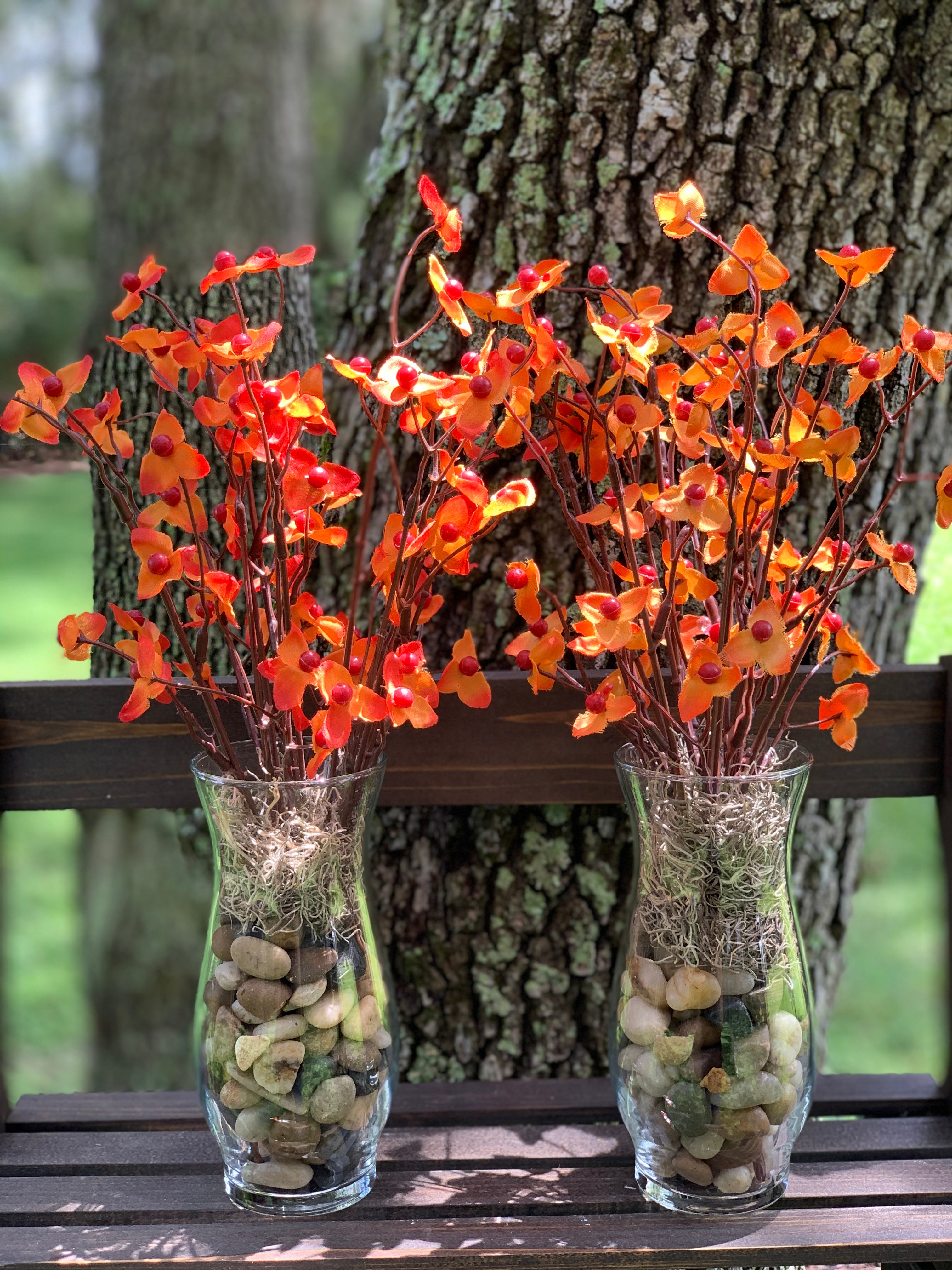Bittersweet Floral Glass Vase shows an image of two vases together.  Each vase has tricolored earth tone rocks and tan moss with orange bittersweet artificial floral decor. Each sold separately.  