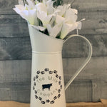 Vintage White Metal Milk Pitcher shows image sitting on a table with a floral arrangement.  Flowers not sold with pitcher.