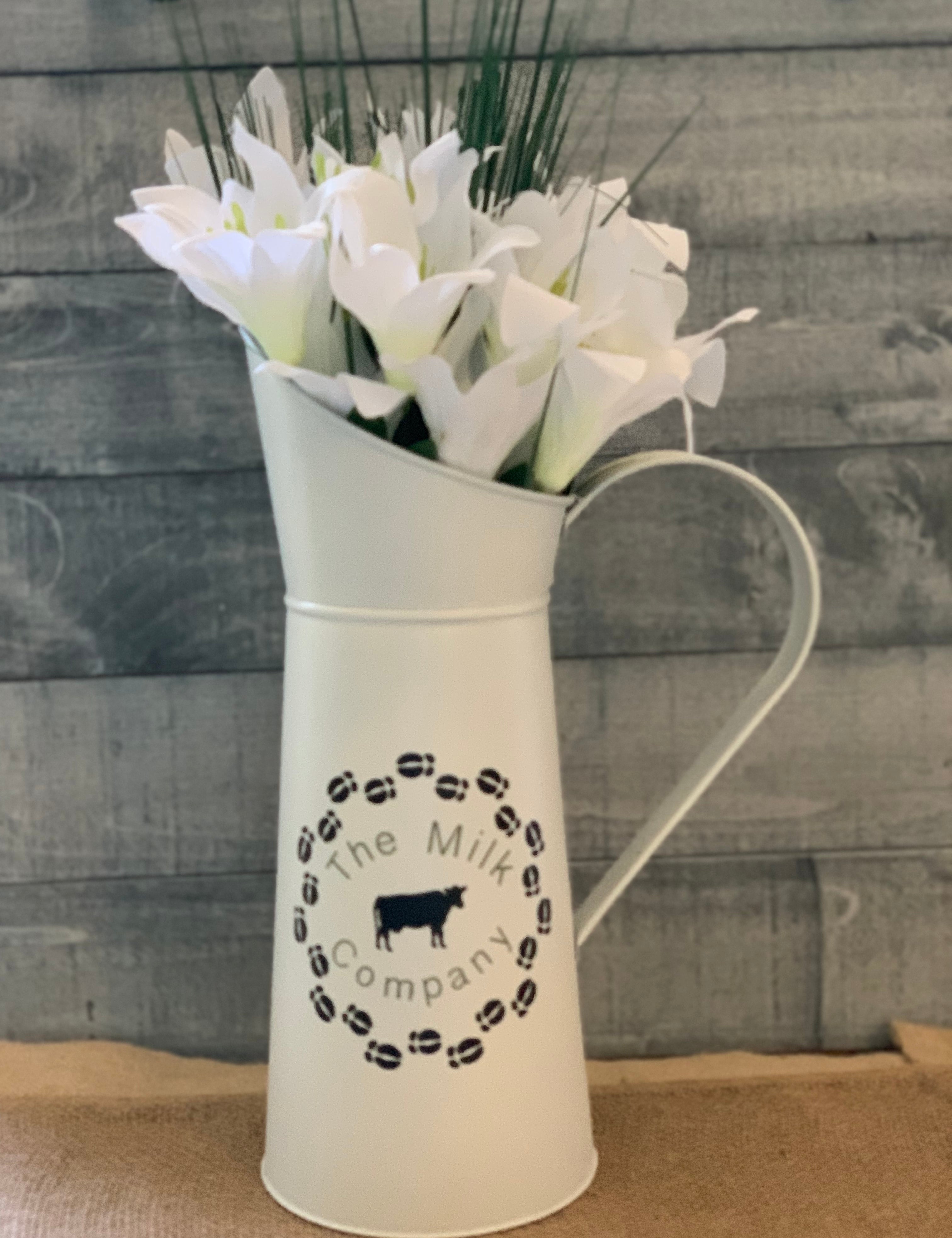 Vintage White Metal Milk Pitcher shows image sitting on a table with a floral arrangement.  Flowers not sold with pitcher.