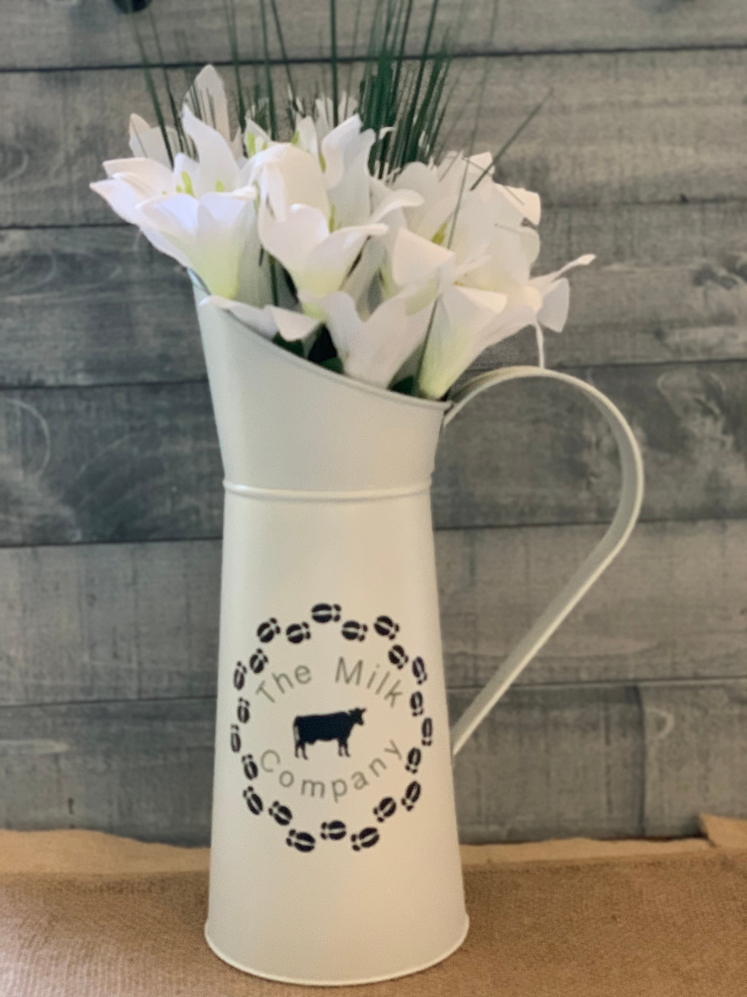Vintage White Metal Milk Pitcher shows image sitting on a table with a floral arrangement.  Flowers not sold with pitcher.