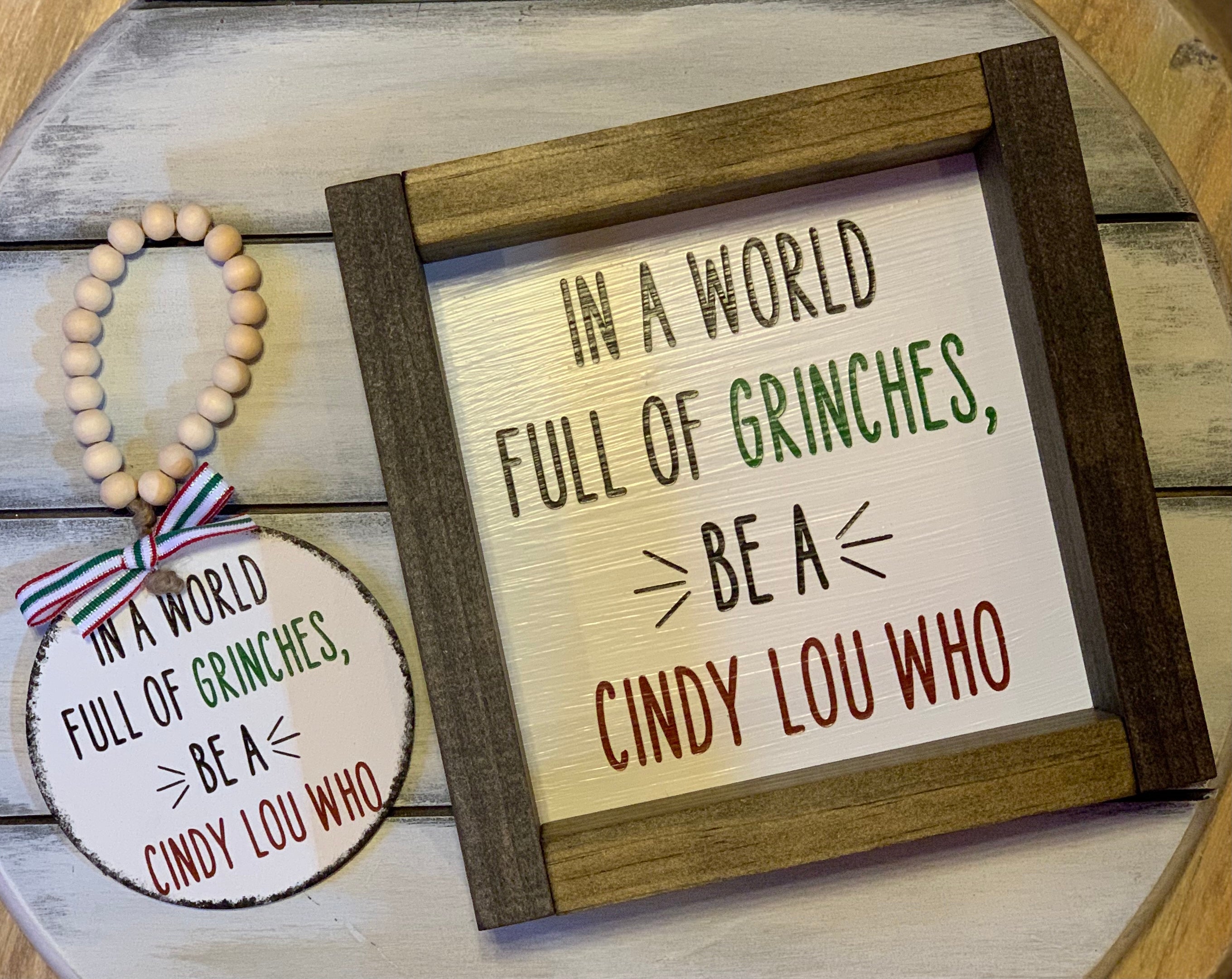 In A World Full of Grinches, Be A Cindy Lou Who 7x7 Sign is shown with the matching ornament.  Each product sold separately.