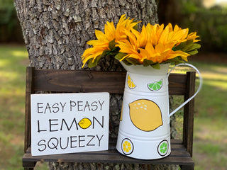 White Metal Lemon and Lime Pitcher shows an image of the pitchers with sunflowers and the easy peasy lemon squeezy sign sitting on a ladder outside. Each item sold separately.