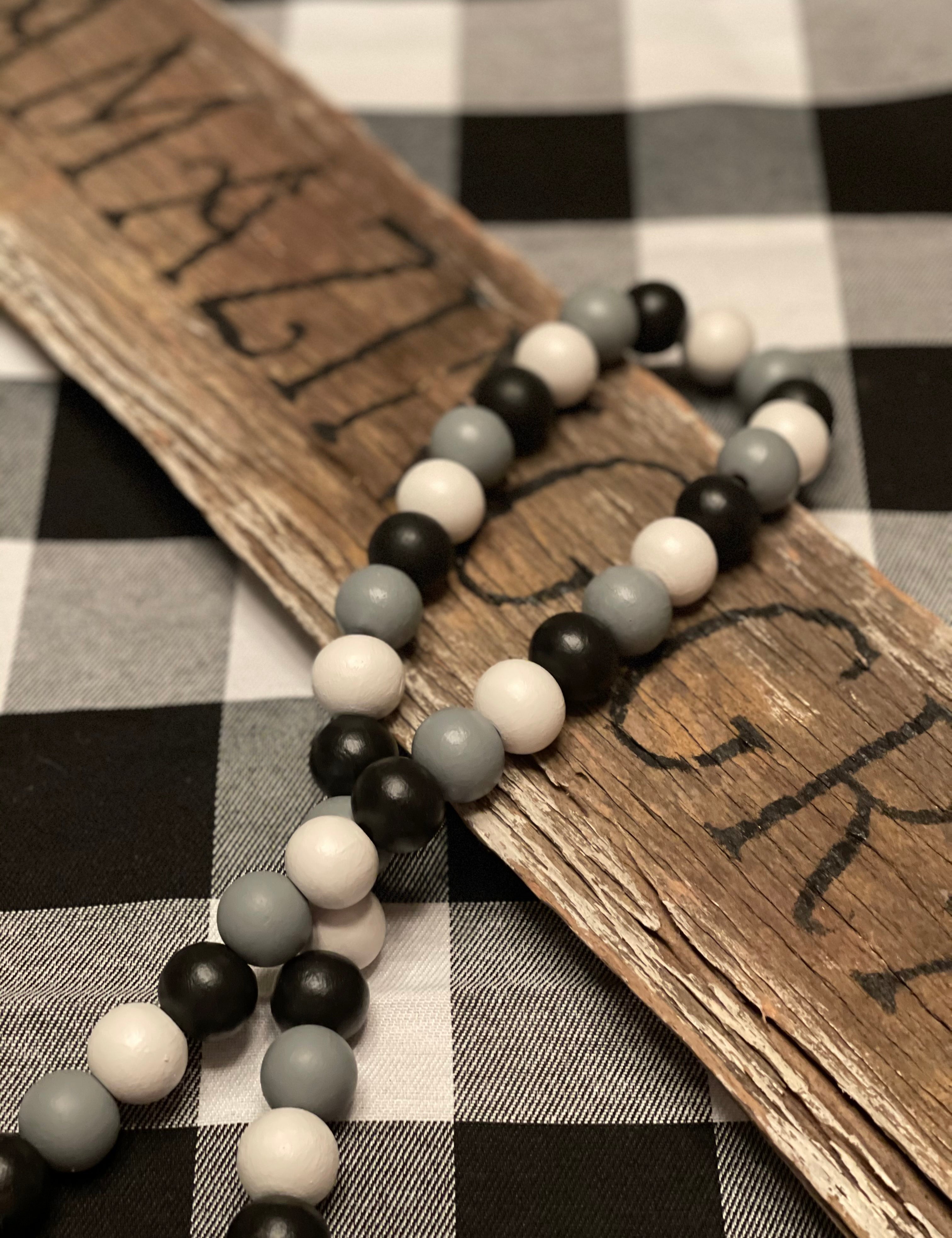 Buffalo Plaid Wood Bead Garland is shown laying across a wood sign.  (Sign is not for sale). The bead colors are white, black and gray with white yarn tassels. 