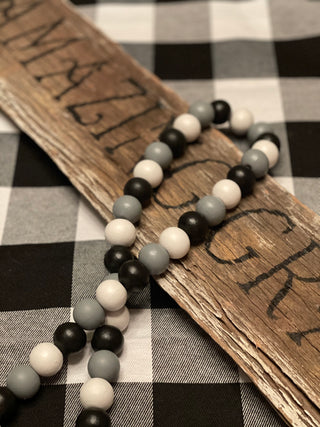 Buffalo Plaid Wood Bead Garland is shown laying across a wood sign.  (Sign is not for sale). The bead colors are white, black and gray with white yarn tassels. 