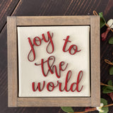 This image of Joy To The World is shown laying on a wood bench with a floral greenery stem.