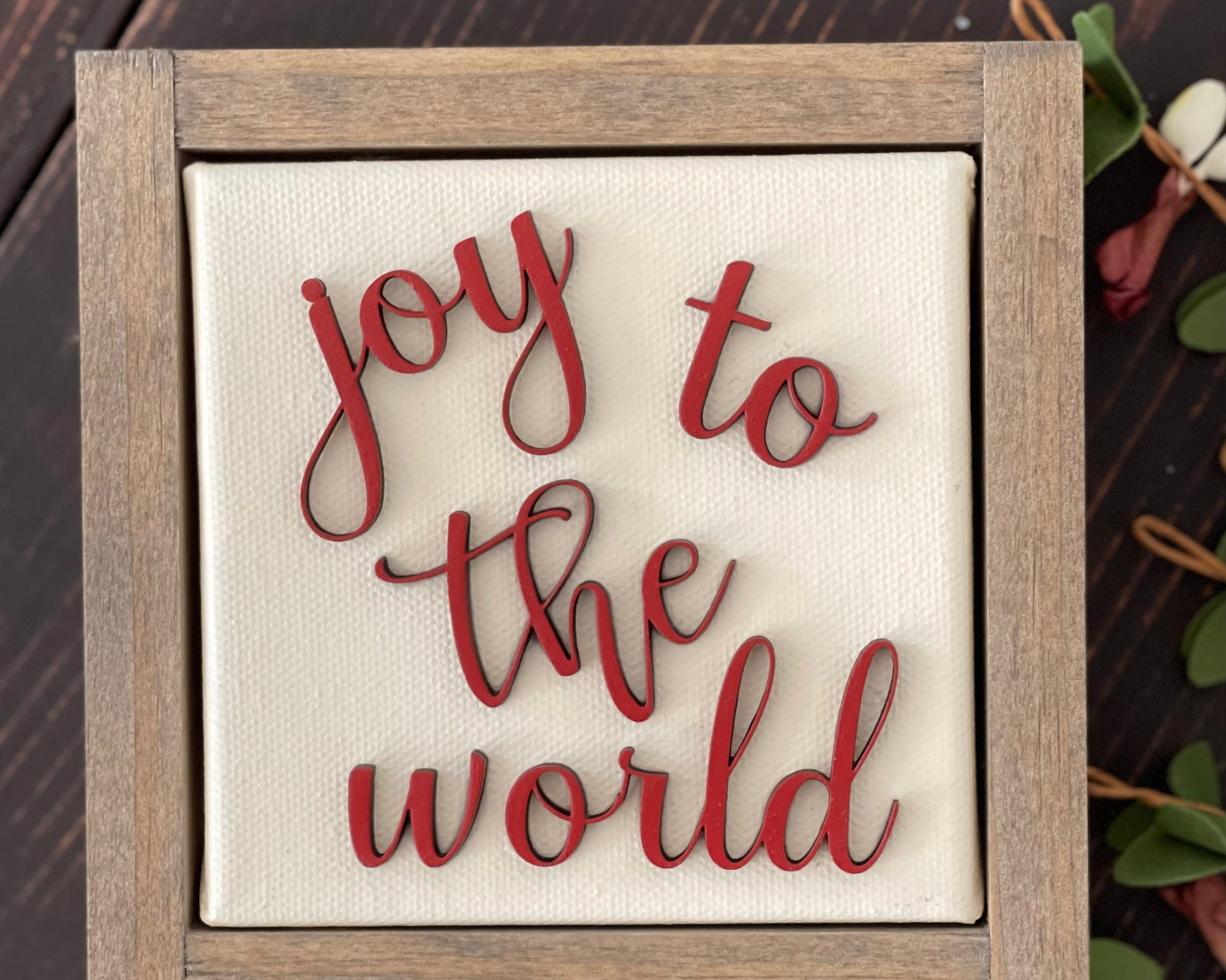 This image of Joy To The World is shown laying on a wood bench with a floral greenery stem.