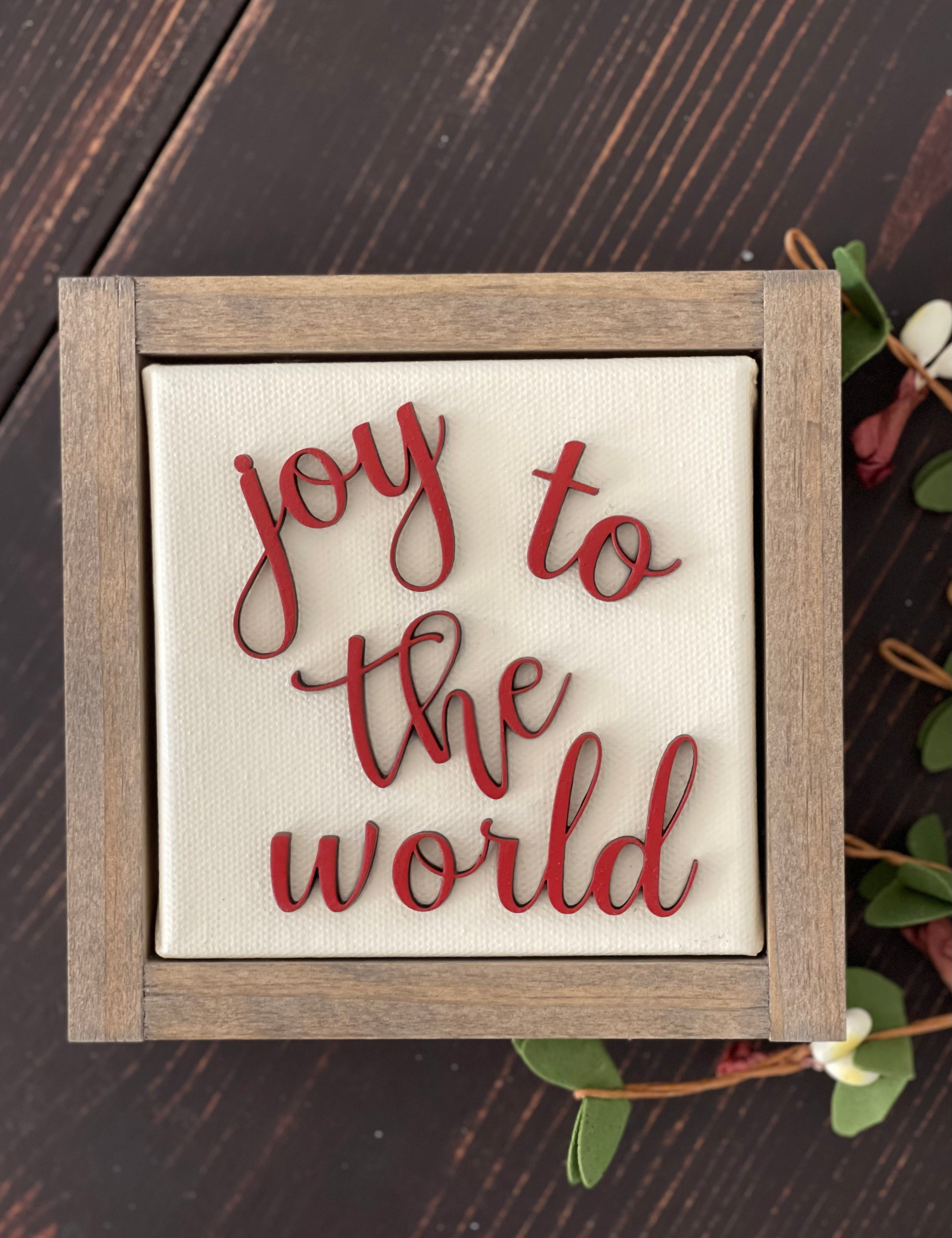 This image of Joy To The World is shown laying on a wood bench with a floral greenery stem.