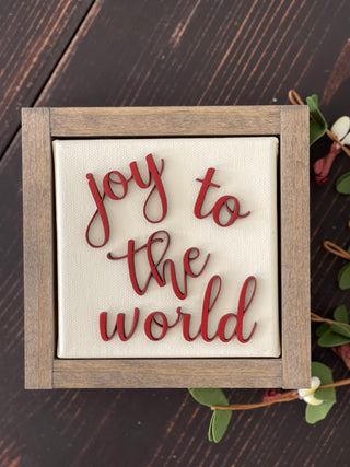 This image of Joy To The World is shown laying on a wood bench with a floral greenery stem.