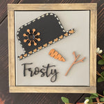 This image shows the Frosty canvas sign laying on a bench with greenery.