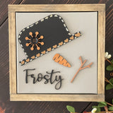 This image shows the Frosty canvas sign laying on a bench with greenery.