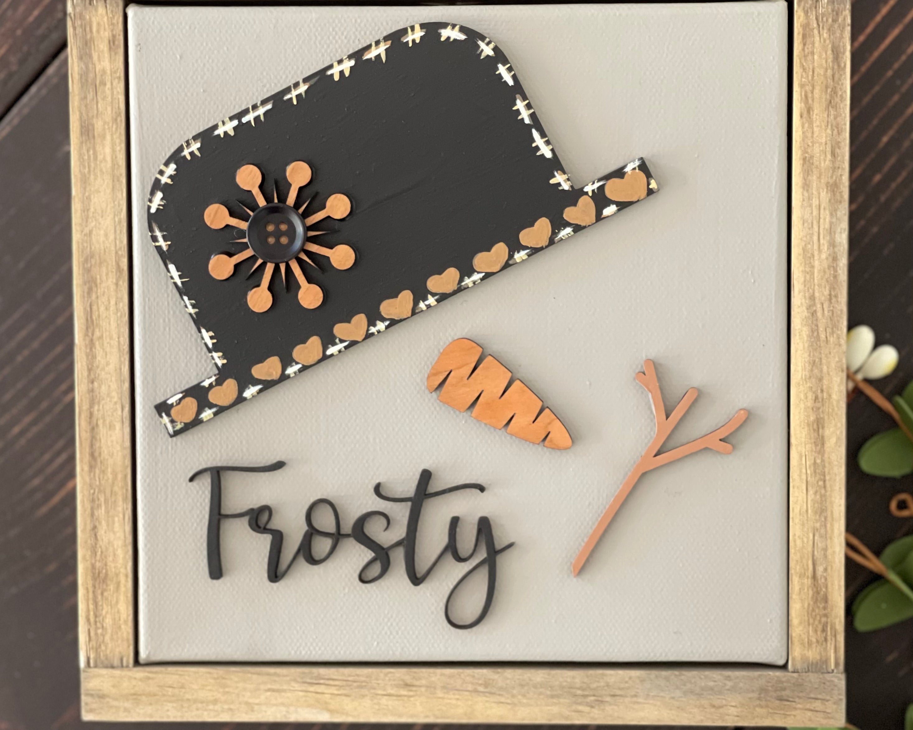This image shows the Frosty canvas sign laying on a bench with greenery.