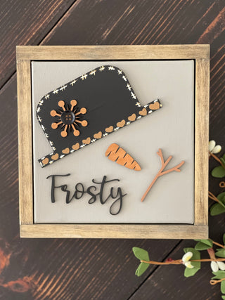 This image shows the Frosty canvas sign laying on a bench with greenery.