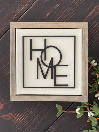 This Home sign is displayed laying on a bench with greenery.
