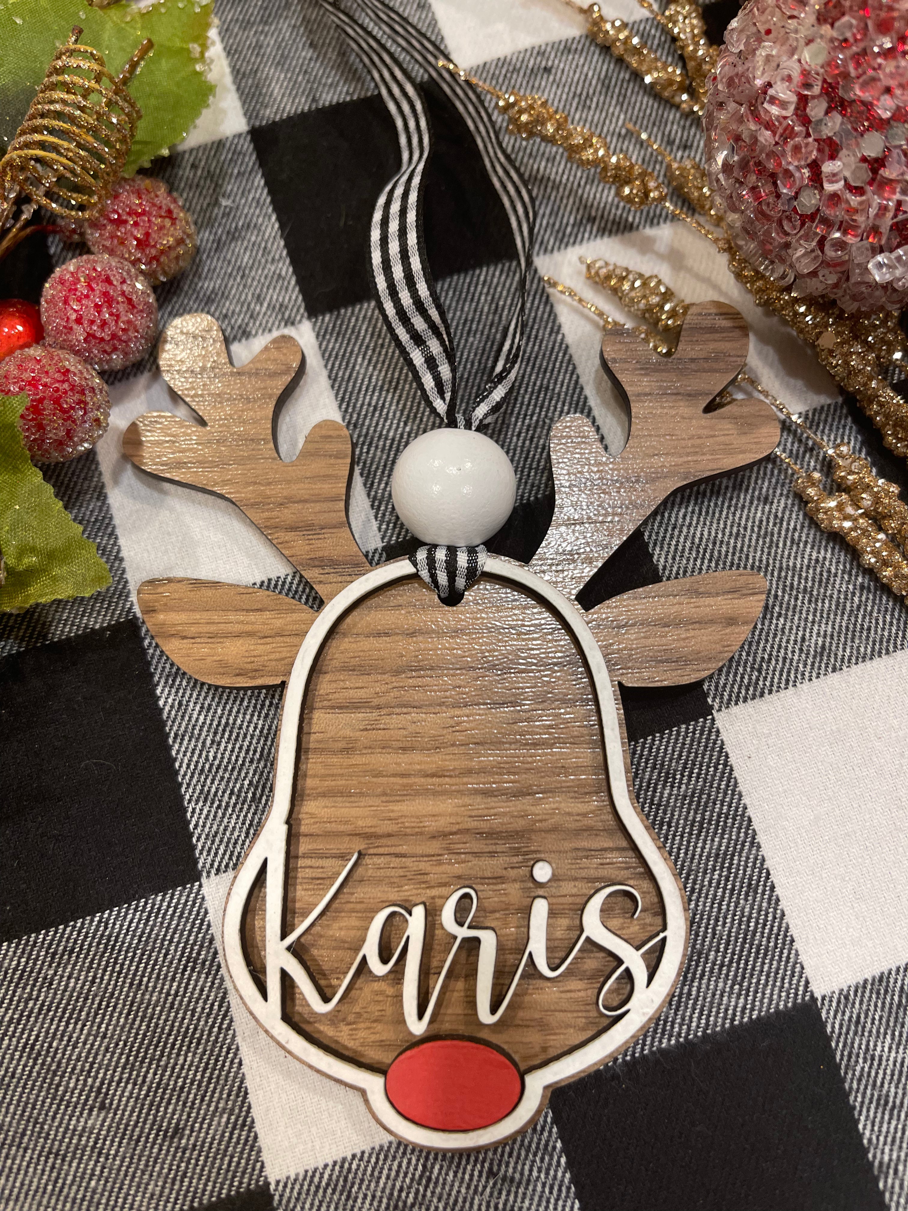 This ornament shows a walnut base with white 3D wood and a name. Buffalo plaid ribbon and a red nose.
