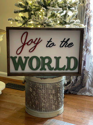 Joy to the world Christmas sign is shown by a flocked Christmas Tree.  Joy is hand painted red, to the is painted black, and world is painted in green.  This sign is a beautiful addition to all your holiday home decor.