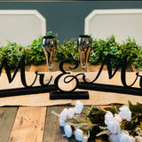 This image shows the black Mr. & Mrs. table topper with greenery and bride and groom toasting glasses.  (All other embellishments not sold with signs).