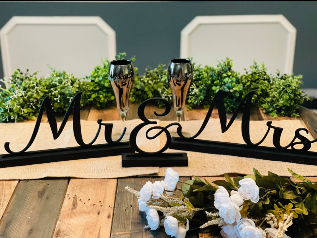 This image shows the black Mr. & Mrs. table topper with greenery and bride and groom toasting glasses.  (All other embellishments not sold with signs).